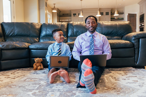 A young boy, dressed up in a shirt and tie, wants to be like his Dad, working from home. The boy works off his homemade cardboard laptop following his role model Dad around, working on business.