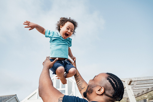 Over the shoulder view of 35 year old Black father lifting ecstatic 4 year old son high into the air as they enjoy outdoor playtime together.