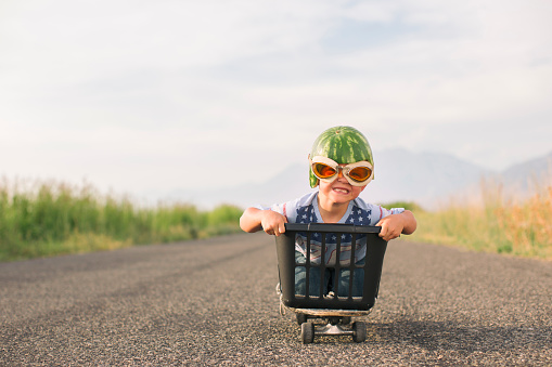 A young boy races his makeshift go-cart while wearing watermelon helmet and goggles. The boy loves speed and smiles as he races down a road.