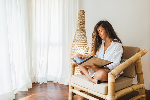 Young brunette woman sitting in wooden bamboo chair indoors and reading book or magazine. Eco style interior concept. Wicker handmade lamp behind.