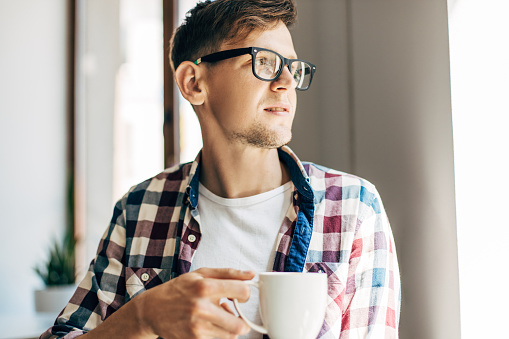 Happy young business man in glasses drinking aromatic coffee from a cup sitting in a cafe near the window