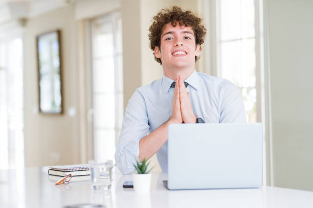 Young business man working with computer laptop at the office praying with hands together asking for forgiveness smiling confident.  prayer request stock pictures, royalty-free photos & images