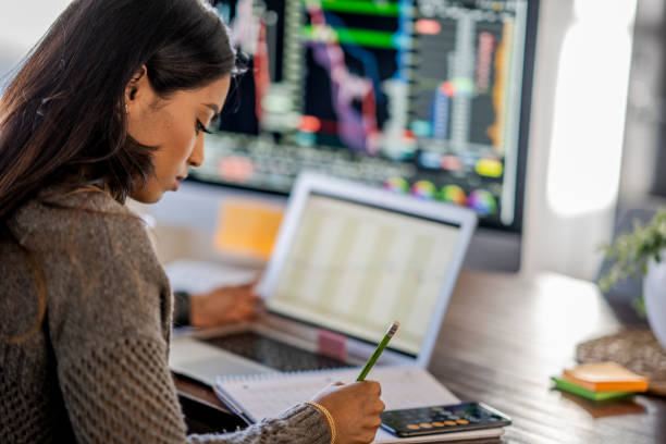 Young business woman day trading from her dining room table Middle Eastern woman tracking and trading stocks using laptop and desktop computer. Stock Exchange, investment, and financial trading concept. data analyst certification stock pictures, royalty-free photos & images