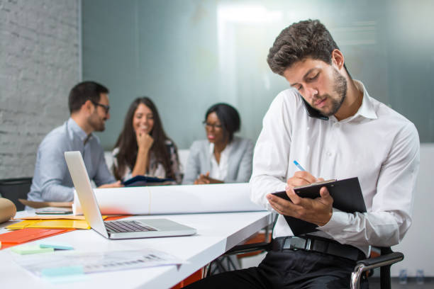 Young businessman holding mobile phone between ear and shoulder talking and writing notes to clipboard in modern office, while his business partners having meeting in the background. stock photo