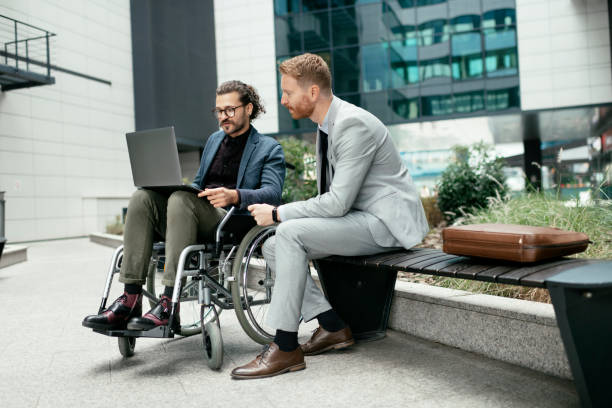A young businessman in a wheelchair is sitting outside with a colleague. stock photo stock photo