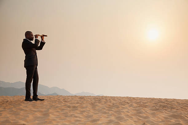 Young businessman looking through telescope in middle of the desert stock photo