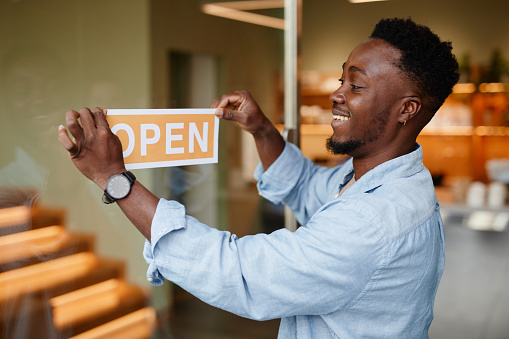 Joyful young African American man opening modern cafe in morning sticking sign on glass door