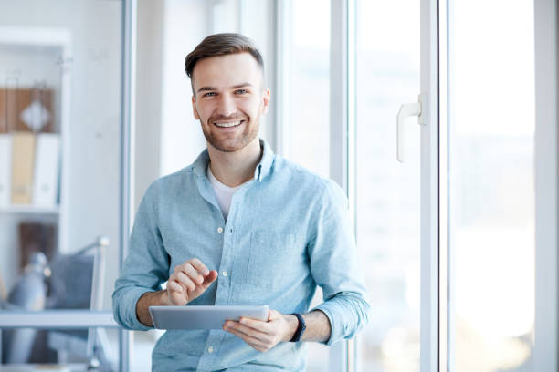 Young Businessman Posing by Window Waist up portrait of handsome young man holding digital tablet and smiling happily at camera standing by window, copy space one young man only stock pictures, royalty-free photos & images