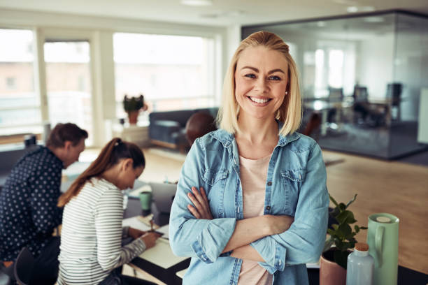 Young businesswoman smiling with coworkers sitting in the background Smiling young businesswoman standing with her arms crossed in an office with colleagues working at a table in the background scandinavia stock pictures, royalty-free photos & images