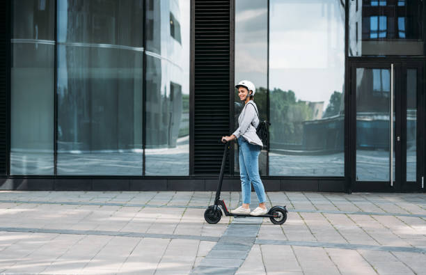 young businesswoman wearing cycling helmet riding push scooter on footpath - elektrische step stockfoto's en -beelden