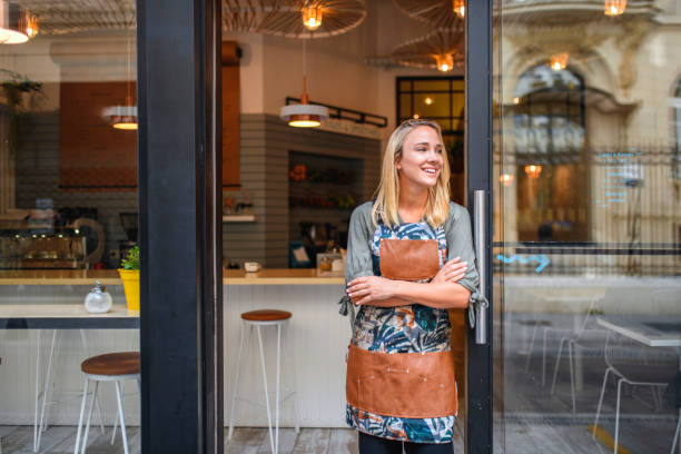 Young Cafe Owner Standing with Arms Crossed at Front Door Cheerful Argentine female cafe owner standing in apron with arms crossed at open front door and looking away from camera. store window stock pictures, royalty-free photos & images