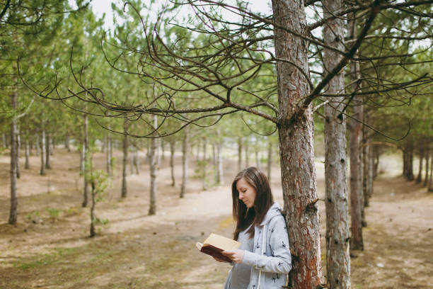 Young calm beautiful woman in casual clothes leaning on tree studying...