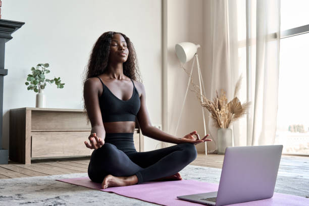 Young calm fit healthy African black woman at home doing yoga learning online class. stock photo