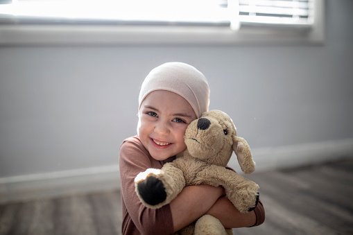 A sweet little girl battling cancer sits at home on the floor of her bedroom, with her arms wrapped tightly around her stuffed animal as she smiles gently for the portrait. She is dressed casually and wearing a headscarf as the sun shines in on her through the window.