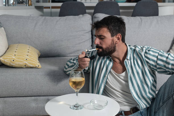 Young carefree man sitting on the floor in his home lighting a cigarette to smoke and drinks wine he has decided not to be unhappy and depressed anymore because life is only one and he wants to live it stock photo