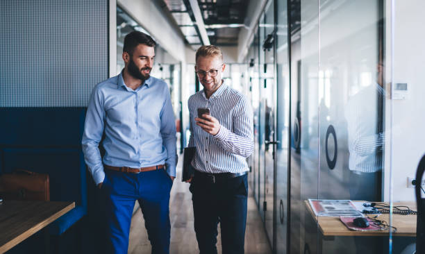 Young caucasian male colleagues having fun on break checking content on mobile phone walking in office, smiling men employees discussing news from exchange web page on free time using smartphone stock photo