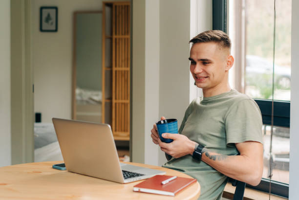 Young caucasian man drinking coffee and using laptop while sitting at home. stock photo