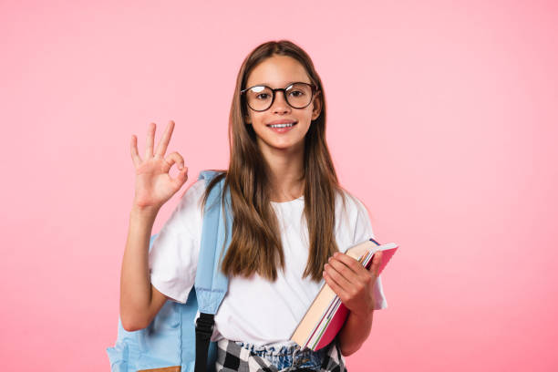 Young caucasian schoolgirl pupil student holding books notebooks showing okay gesture looking at camera isolated in pink background. stock photo