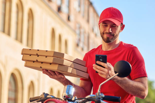 Young cheerful caucasian delivery man in red uniform sitting on scooter and using smartphone while delivering pizza stock photo