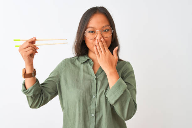 Young chinese woman wearing glasses holding chopsticks over isolated white background cover mouth with hand shocked with shame for mistake, expression of fear, scared in silence, secret concept Young chinese woman wearing glasses holding chopsticks over isolated white background cover mouth with hand shocked with shame for mistake, expression of fear, scared in silence, secret concept how do you say shut up in japanese stock pictures, royalty-free photos & images