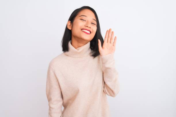 Young chinese woman wearing turtleneck sweater standing over isolated white background Waiving saying hello happy and smiling, friendly welcome gesture  wave goodbye asian stock pictures, royalty-free photos & images