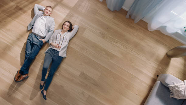 Young Couple are Lying on a Wooden Flooring in an Apartment. They are Happy, Smile and Laugh. Cozy Living Room with Modern Interior, Grey Sofa and Wooden Parquet. Top View Camera Shot. Young Couple are Lying on a Wooden Flooring in an Apartment. They are Happy, Smile and Laugh. Cozy Living Room with Modern Interior, Grey Sofa and Wooden Parquet. Top View Camera Shot. parquet floor stock pictures, royalty-free photos & images