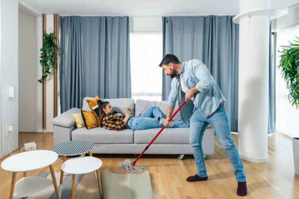 Young couple at home a man wipes and cleaning the floor so his woman can rest and browses the internet on her smartphone and lying on the sofa so that he can clean the floor from dust and dirt stock photo