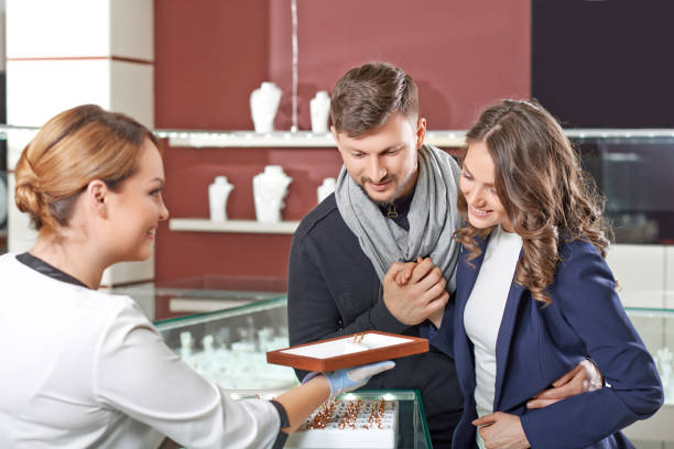 Young couple buying jewelry Timeless design. Friendly female jeweler helping young engaged couple to choose wedding rings store clerk selling jewelry stock pictures, royalty-free photos & images