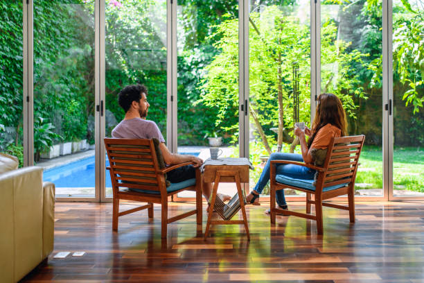 Pareja joven tomando café matutino en casa en la sala de estar Vista trasera de una mujer hispana y un hombre de raza mixta de 20 y 30 años sentados con café en una sala de estar con luz natural con vista al patio trasero y a la piscina. mesa de cafe fotos e imágenes de stock