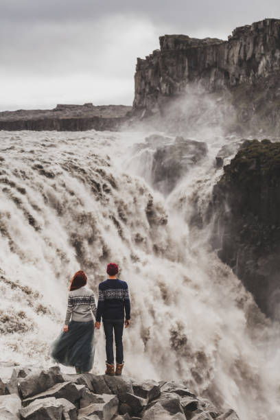 Young couple in love looking at famous icelandic landmark Dettifoss waterfall. Traditional wool sweaters, red hair, hat, gray skirt. Dramatic nordic landscape, cold weather in Iceland. View from behind. Young couple in love looking at famous icelandic landmark Dettifoss waterfall. Traditional wool sweaters, red hair, hat, gray skirt. Dramatic nordic landscape, cold weather in Iceland. View from behind. iceland dettifoss stock pictures, royalty-free photos & images