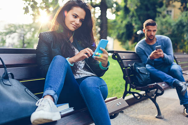 Young couple in the park texting on smartphones Teenage couple using smartphones in the city park flirting stock pictures, royalty-free photos & images