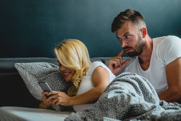 Young couple is having relationship problems Youg men is watching his girlfriends phone over her sholder while they are laying in bed envy stock pictures, royalty-free photos & images