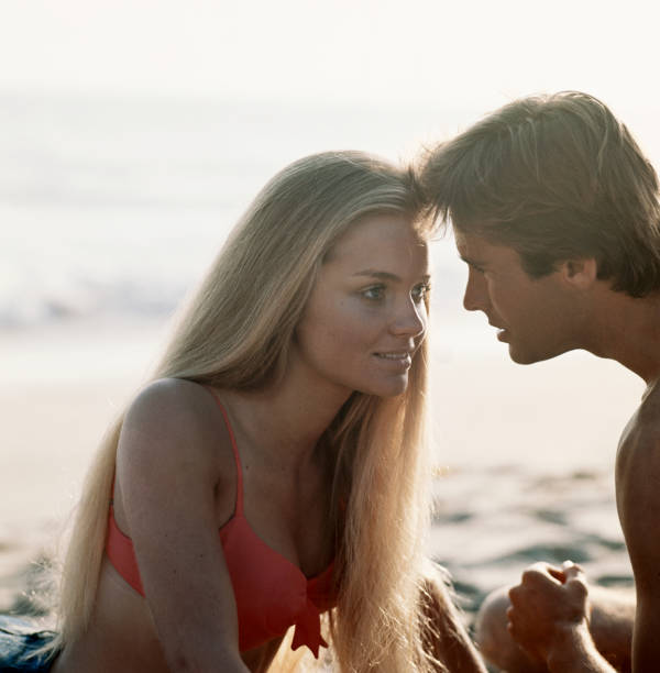 young couple looking at each other on beach - man strand blond haar jaren zeventig stockfoto's en -beelden
