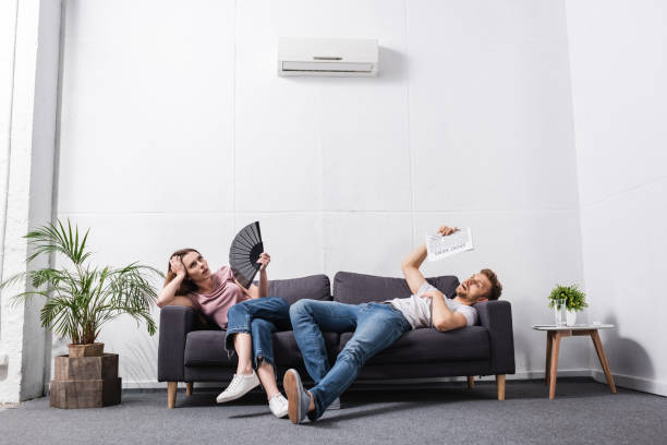 young couple with hand fan and newspaper suffering from heat at home with broken air conditioner young couple with hand fan and newspaper suffering from heat at home with broken air conditioner broken stock pictures, royalty-free photos & images