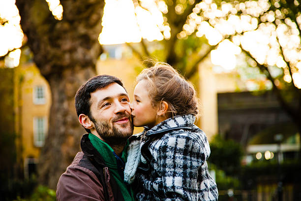 A young girl kisses her her father in an autumn park while she is kept in his arms.