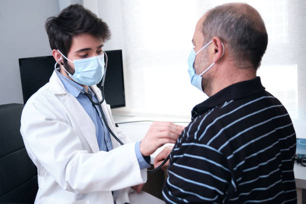 Young doctor health care provider performing a physical exam and listening to mature man patient heart and lungs with a stethoscope. stock photo