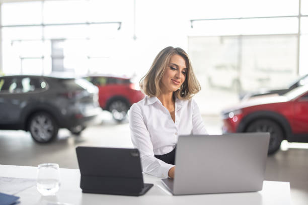 Young elegant sales woman sitting in car dealership saloon and using laptop Young well dressed sales woman sitting at desk in car dealership saloon and using laptop car salesperson stock pictures, royalty-free photos & images