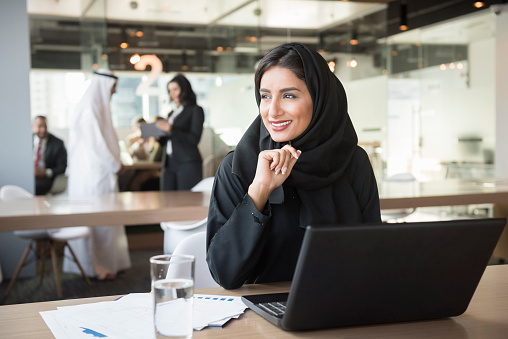 A photo of smiling Emirati businesswoman with laptop. Middle Eastern professional in traditional arab attire of abaya and hijab. She is sitting at conference table. Office worker is looking away while working in brightly lit office.Dubai, United Arab Emirates.