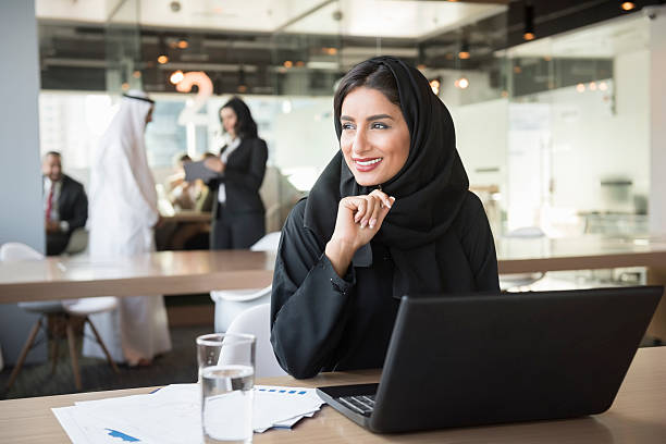 Young Emirati businesswoman looking away at conference table A photo of smiling Emirati businesswoman with laptop. Middle Eastern professional in traditional arab attire of abaya and hijab. She is sitting at conference table. Office worker is looking away while working in brightly lit office.Dubai, United Arab Emirates. abaya clothing stock pictures, royalty-free photos & images