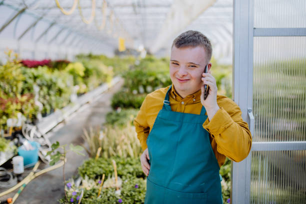 Young employee with Down syndrome working in garden centre, standing in door of greenhouse and calling on cellphone. stock photo