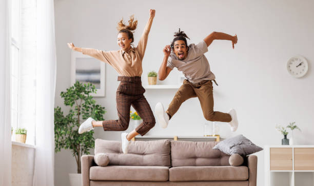 Young energetic active african american family couple jumping on sofa, having fun together at home stock photo