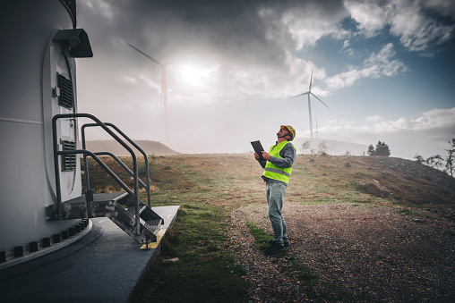Portrait of young caucasian technician engineer man wearing yellow worker headwear standing, working, checking wind farm field system and looking up verify at wind turbine while using a radio station energy plan by wind turbine power generation station