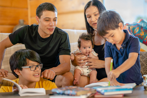 A photo of a young Indigenous Canadian family spending time together in the living room at home. The family consists of a mother, father and their three young children. There are some books open on the coffee table. The two older children are reading and writing on the books.
