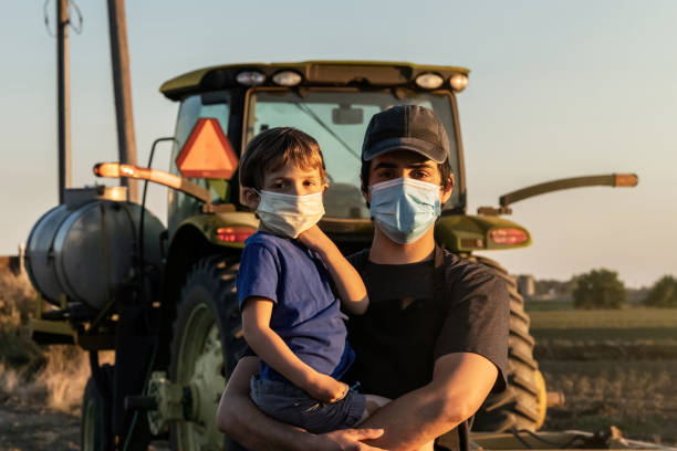 jonge landbouwer die met zijn zoon stelt, allebei die beschermende gezichtsmaskers dragen - landelijke scène stockfoto's en -beelden