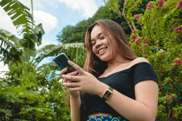A young fashionable asian teen enjoys chatting with her friend or boyfriend and wears a pretty smile. Wearing a black off-shoulder blouse and holding a blue smartphone. Tropical garden setting. stock photo
