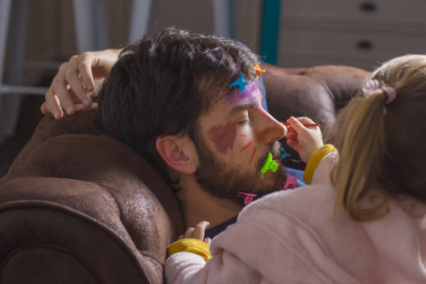 Young father sleeping on the couch while his little daughter paints his face with colorful watercolors Father's Day celebration. Portrait of young father sleeping on the sofa while his little daughter paints her face with colorful watercolors and puts hair clips on her beard child behaving badly stock pictures, royalty-free photos & images