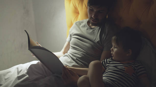 Young father with little son lying on bed at home and reading fairytale book before sleeping in evening stock photo