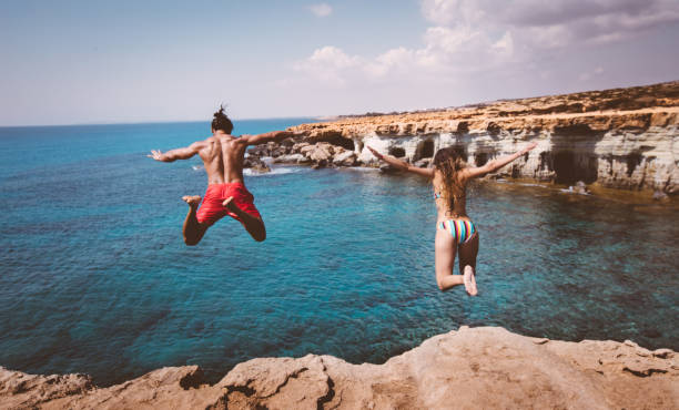 Young fearless divers couple jumping off cliff into ocean Young brave boyfriend and girlfriend jumping off cliff and diving into blue sea water cliff jumping stock pictures, royalty-free photos & images
