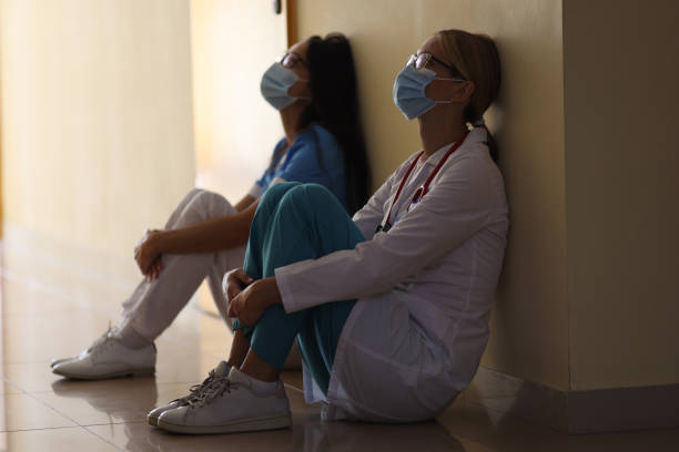 Young female doctor and nurse in medical masks sit in corridor of hospital Young female doctor and nurse in medical masks sit in corridor of hospital. Emotional burnout of healthcare workers during covid19 pandemic concept. mental burnout stock pictures, royalty-free photos & images