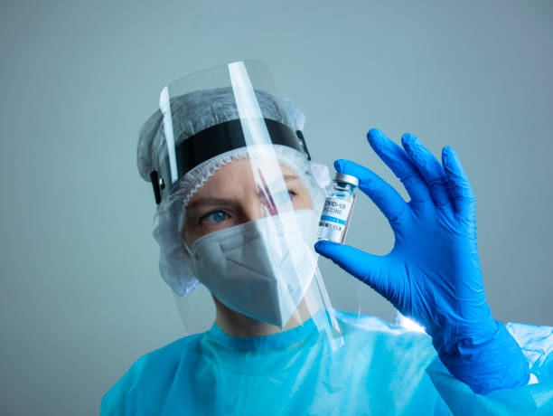young female doctor looks at one bottle of vaccine, nurse wears protective mask and protective suit while holding the vaccine stock photo